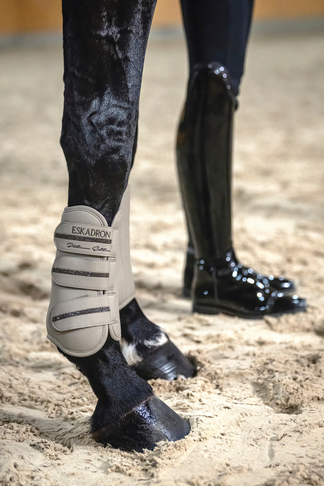 Side view of a black horse wearing the Pale Grey Eskadron Allround Front Faux Leather Tendon Boots while standing in arena footing