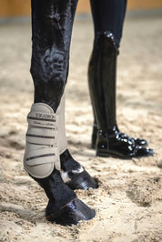 Side view of a black horse wearing the Pale Grey Eskadron Allround Front Faux Leather Tendon Boots while standing in arena footing