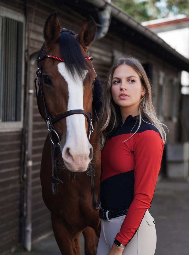 Front view of a bay horse and a female model who is wearing the black/red Criniere Sophie Fierce Focus Long Sleeve Shirt with a barn in the background