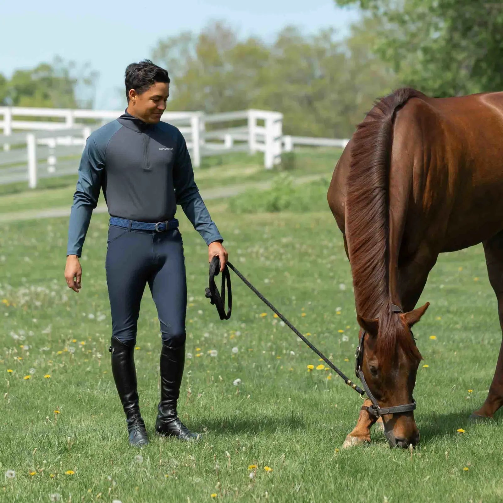 Front view of a man grazing a horse while wearing the navy Equinavia Karl Men's Knee Patch Breeches, matching shirt, and black tall boots