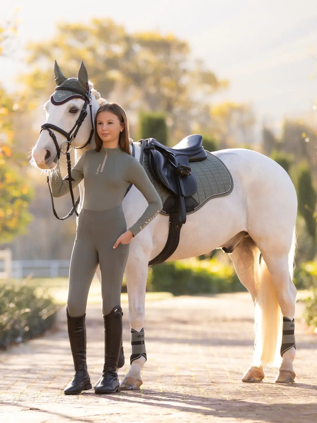 Full body front view of a young female model wearing the green Lemieux Young Rider Orla Pull On Tight and coordinating green top while standing in front of a white pony