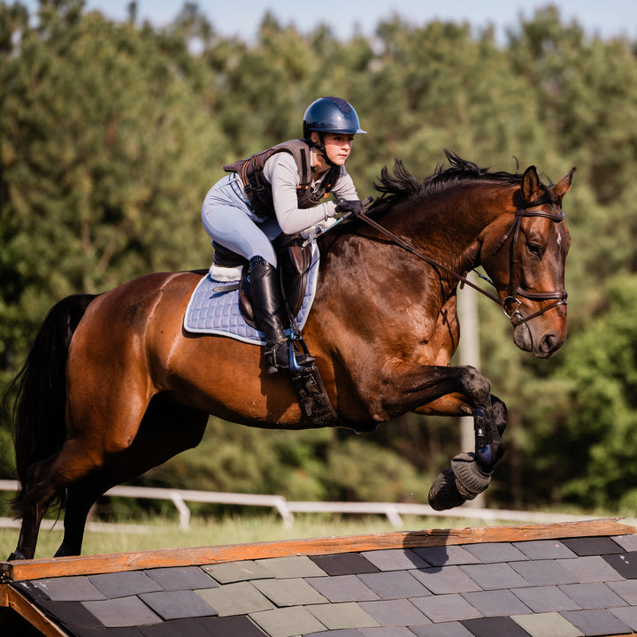 Side view of a model wearing the Navy/Navy Shimmer Equinavia Crown Glossy MIPS Riding Helmet while riding a horse over a jump