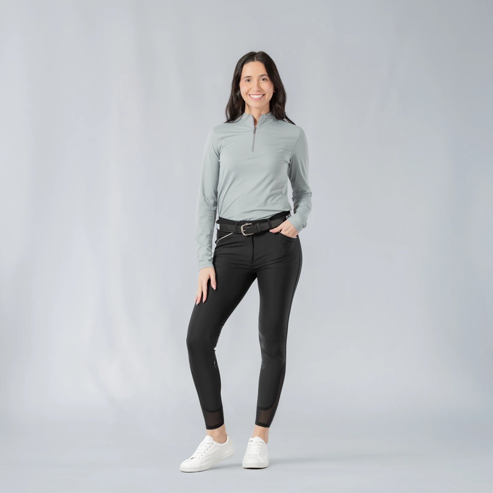 Full body front view of a model wearing the Equinavia Kari Womens Long Sleeve Sun Shirt with NordicAir Tech in gray, black breeches, and white sneakers on a gray background