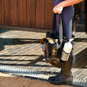 View of a model standing in a barn aisle while holding the Hairy Pony Horse Wash Bay Bag that is filled with bathing supplies