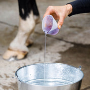 View of someone pouring Hairy Pony Essential Oil Coat Conditioner from a cup into a metal bucket