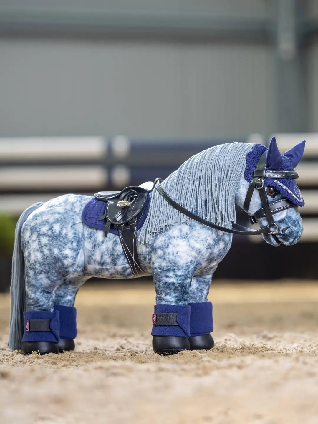 Side view of a gray toy pony wearing the navy LeMieux Toy Pony Saddle Pad and matching set while standing in an indoor arena