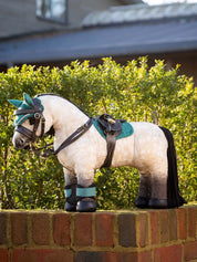 Side view of a buckskin toy pony standing on a brick wall wearing the brown LeMieux Toy Pony Saddle and Girth and green accessories