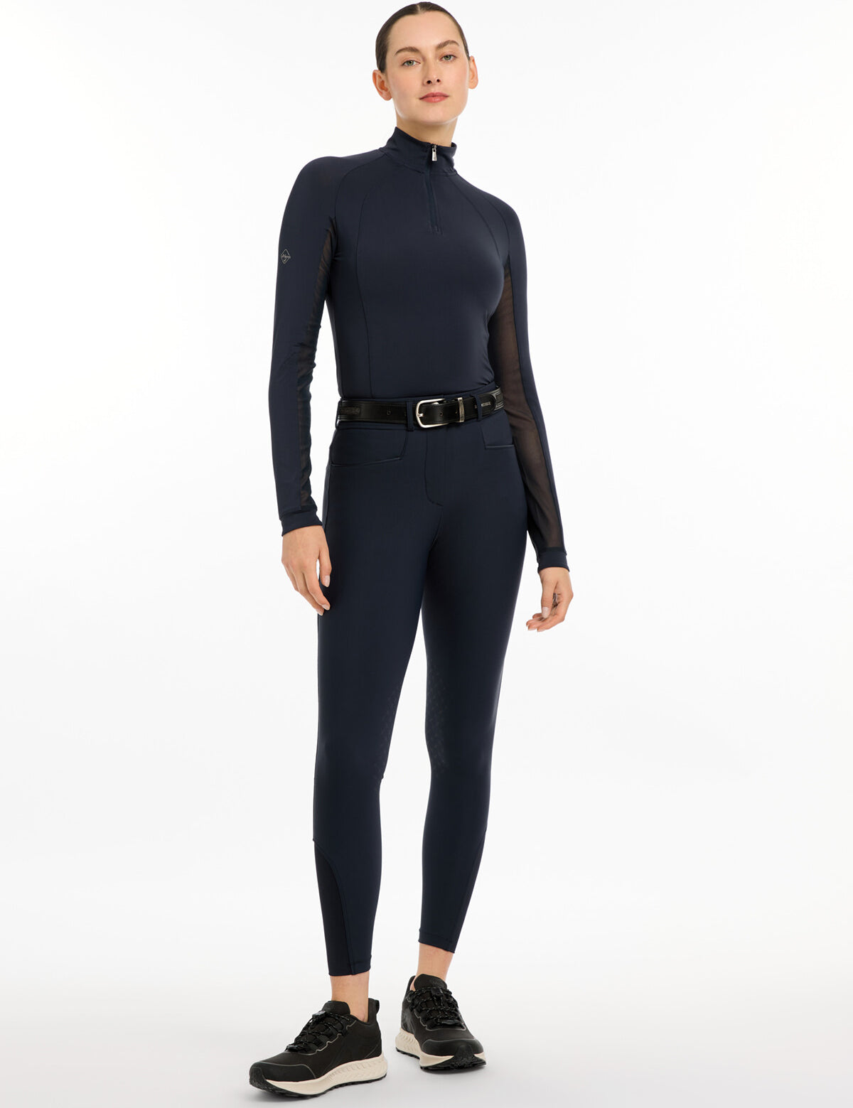 Full body front view of a model wearing the navy LeMieux Isabelle Knee Grip Breeches, navy top, and black shoes on a white background