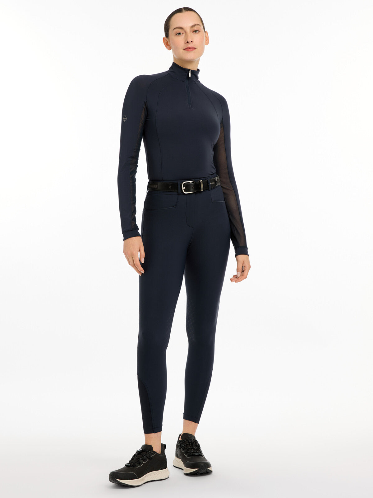 Full body front view of a model wearing the navy LeMieux Isabelle Knee Grip Breeches, navy top, and black shoes on a white background