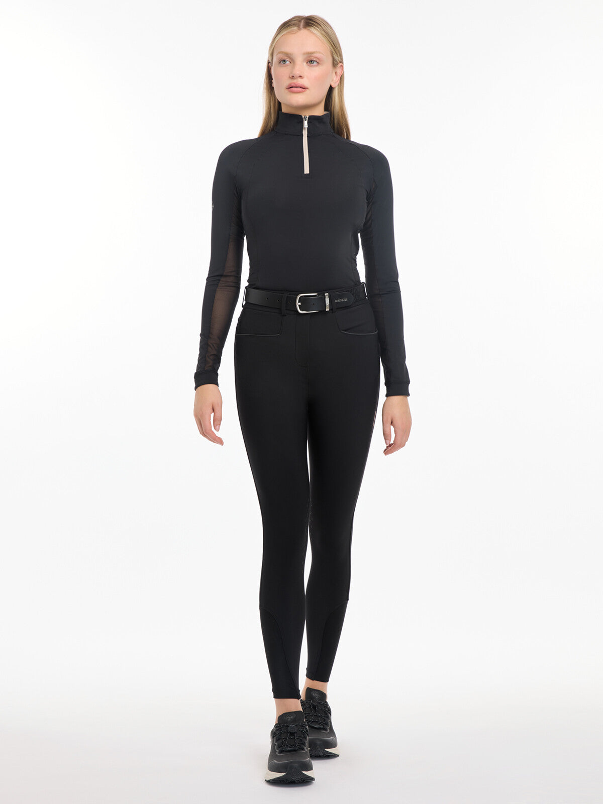 Full body front view of a female model wearing the black LeMieux Isabelle Knee Grip Breeches and black top on a white background