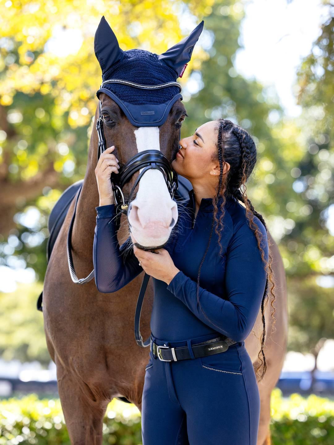 Front view of a female model cuddling the face of a brown horse that is wearing the navy LeMieux Essence Acoustic Fly Hood