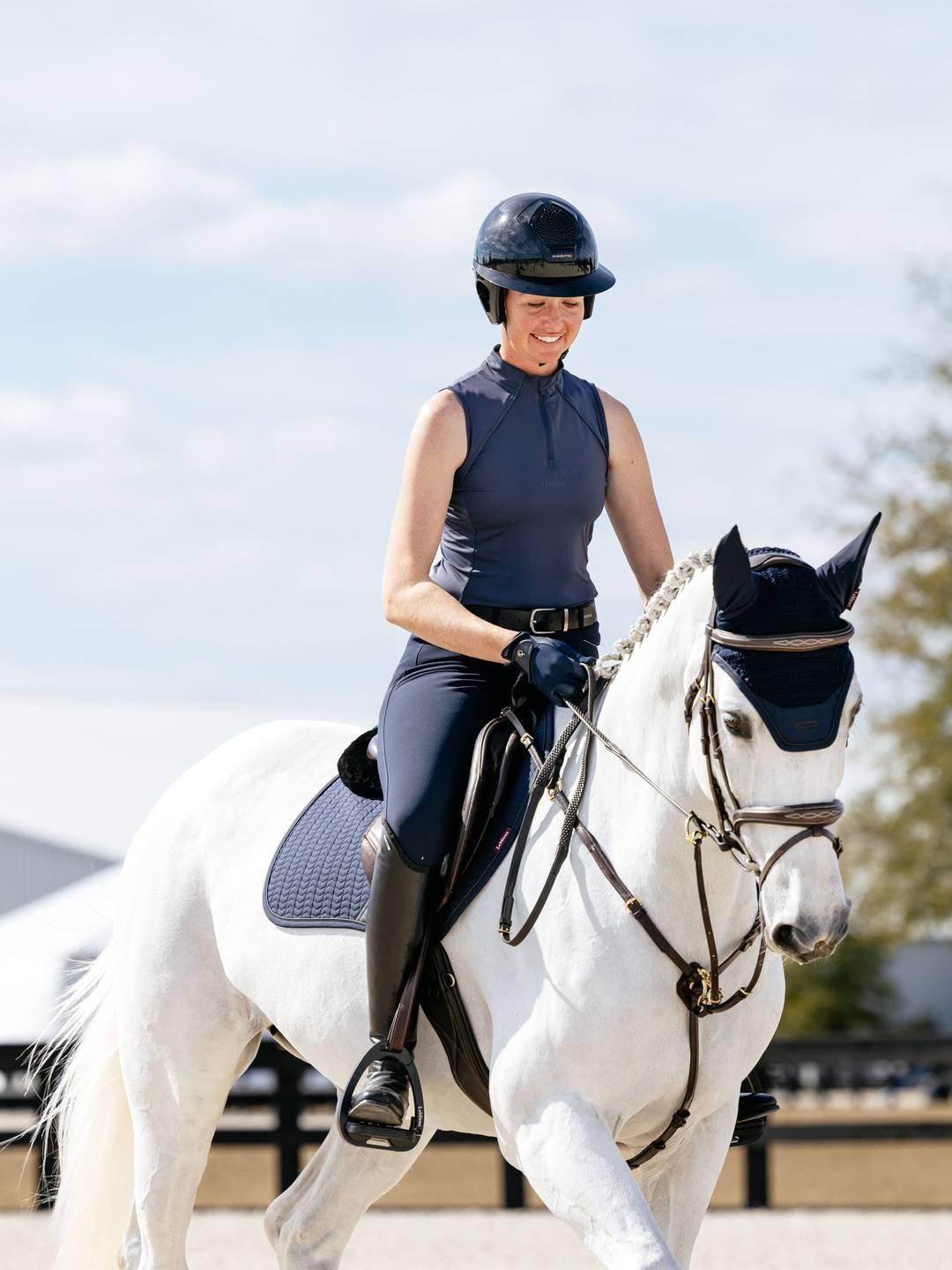 Front view of a female model riding a white horse that is wearing the navy LeMieux Essence Acoustic Fly Hood