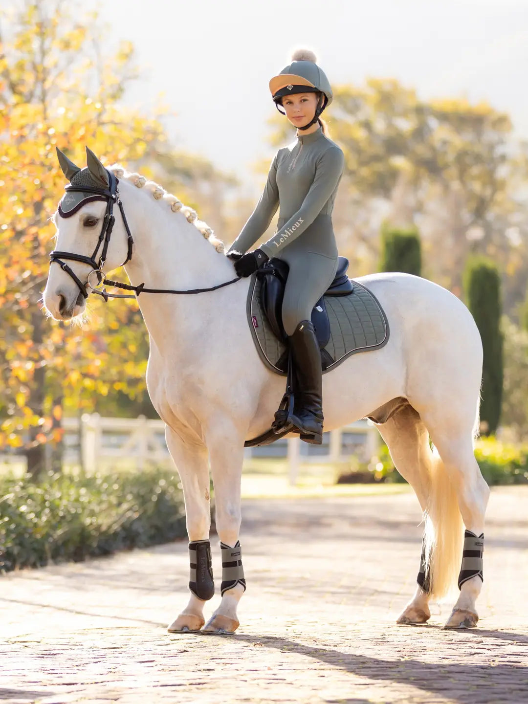 Angled front view of a young female model wearing the green Lemieux Young Rider Orla Pull On Tight and coordinating green top while riding a white pony