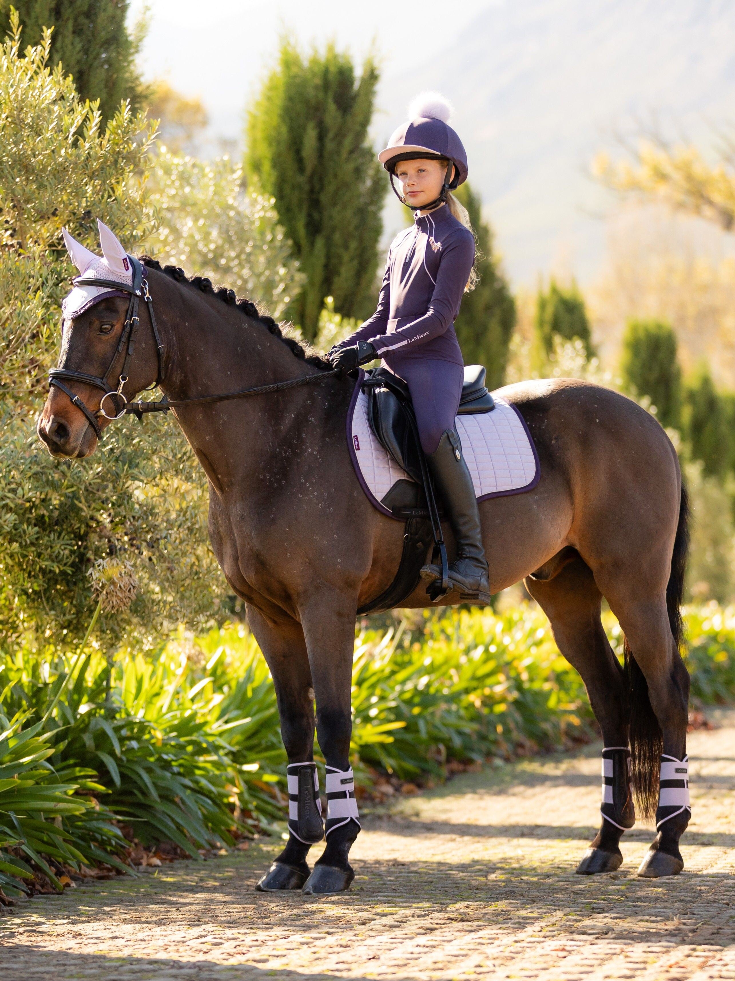 Angled front view of a young female model wearing the purple Lemieux Young Rider Orla Pull On Tight and coordinating purple top while riding a pony
