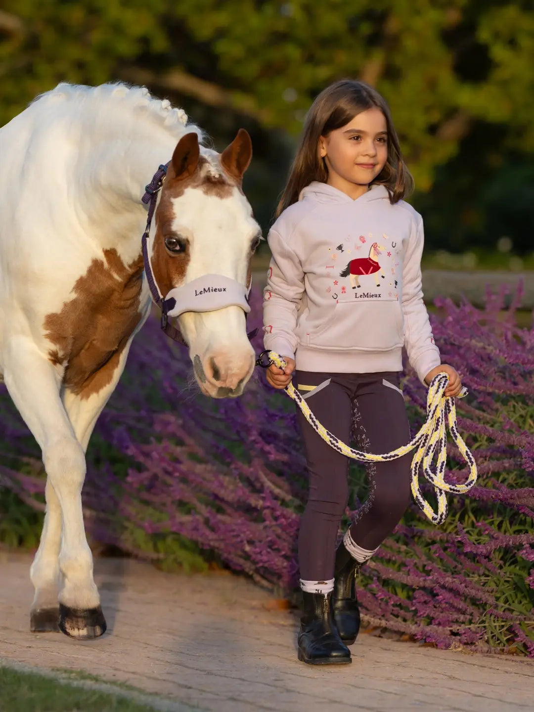 Front view of a child wearing the purple LeMieux Mini Romie Hoody while leading a paint pony outside