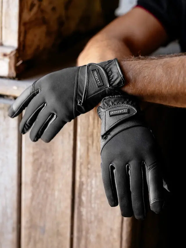 Black insulated gloves worn on a person's hands, shown against a rustic wooden background. The gloves feature a textured knit body, smooth synthetic finger panels, and a branded wrist strap patch.