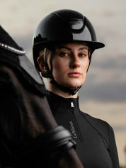 Angled front view of a female model wearing the black LeMieux Verv® Competition Gloss Helmet while standing behind a dark colored horse