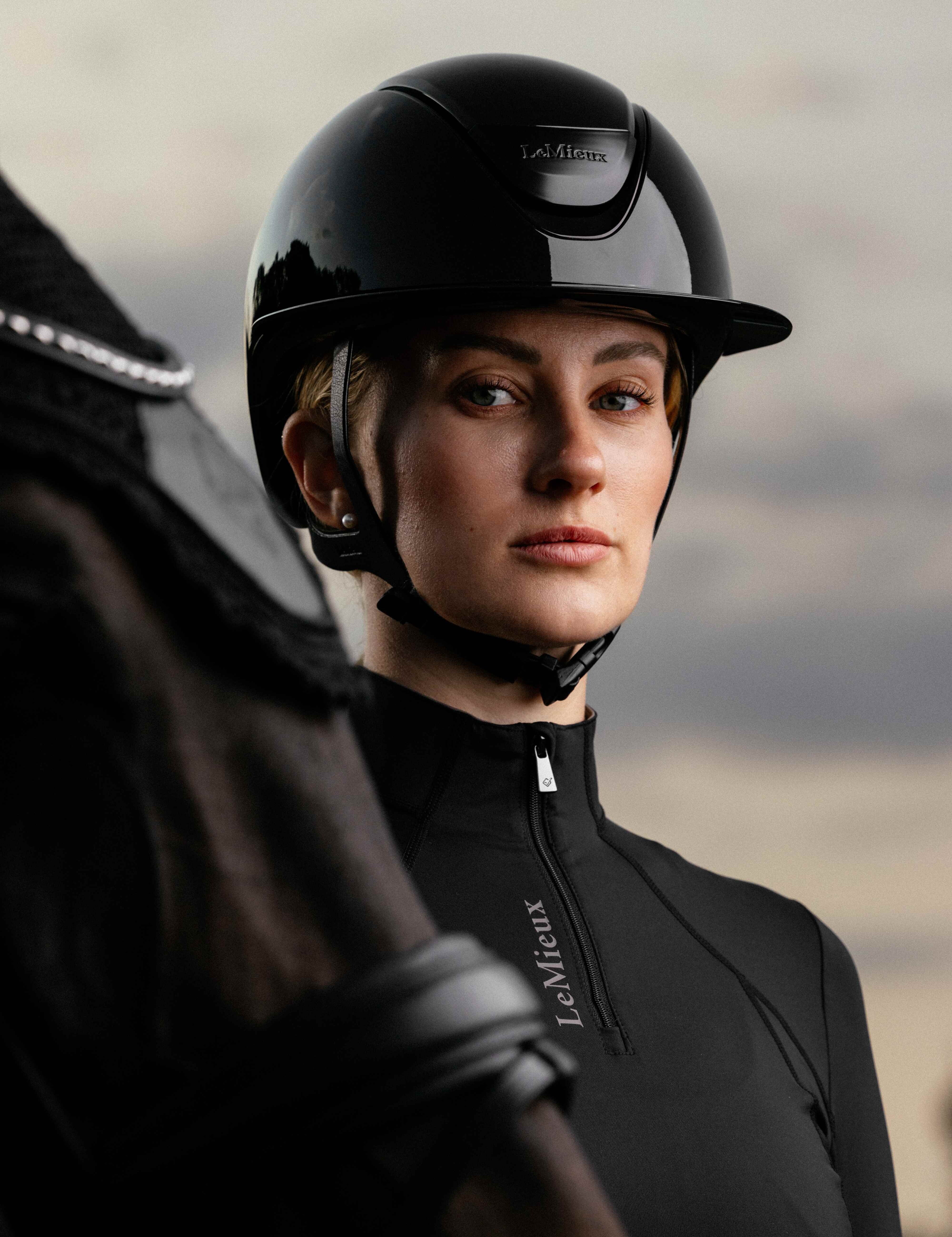Angled front view of a female model wearing the black LeMieux Verv® Competition Gloss Helmet while standing behind a dark colored horse
