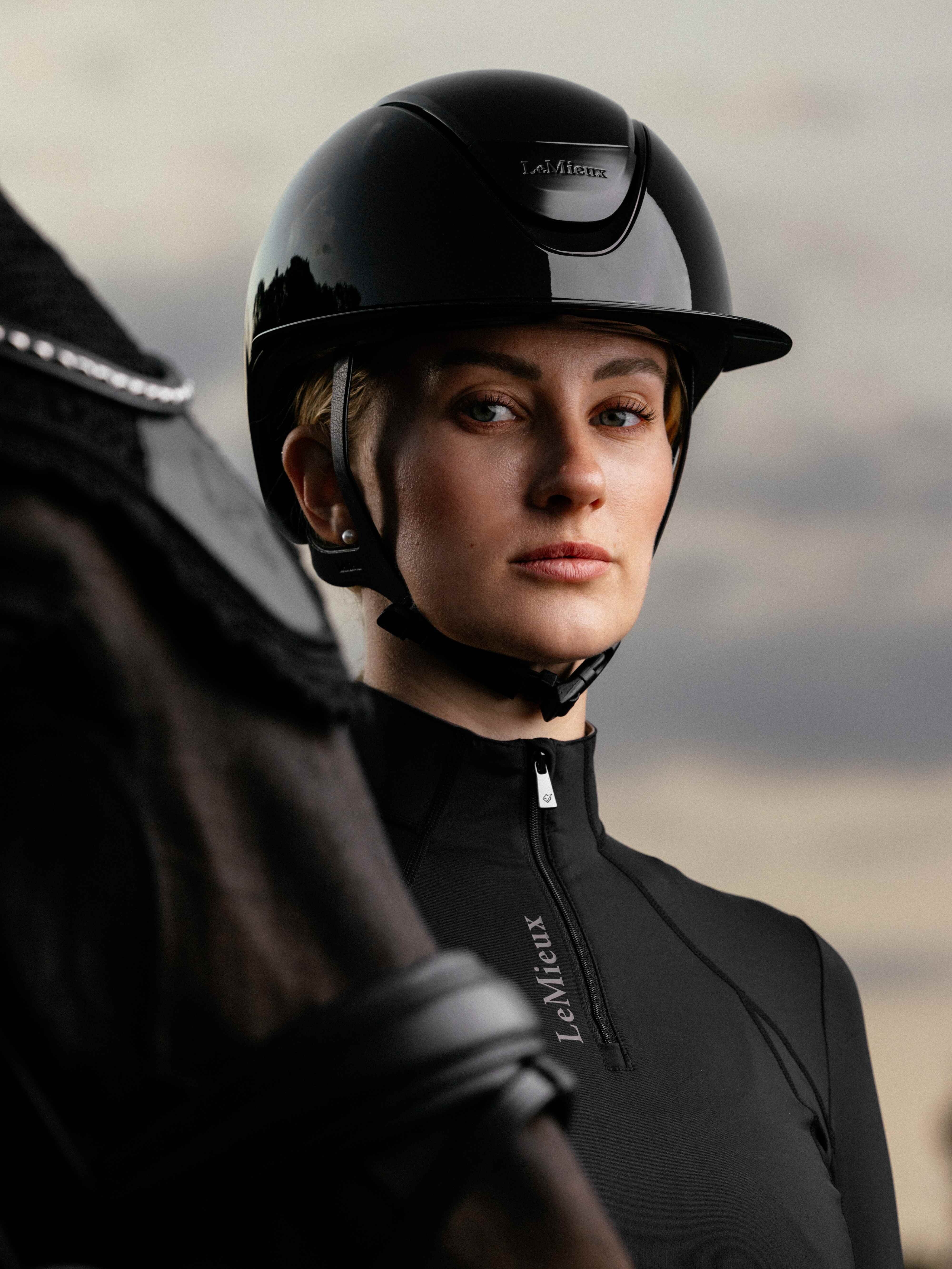 Angled front view of a female model wearing the black LeMieux Verv® Competition Gloss Helmet while standing behind a dark colored horse