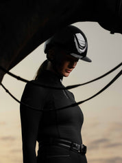 Side view of a female model wearing the black LeMieux Verv® Competition Gloss Helmet while standing behind a horse's neck