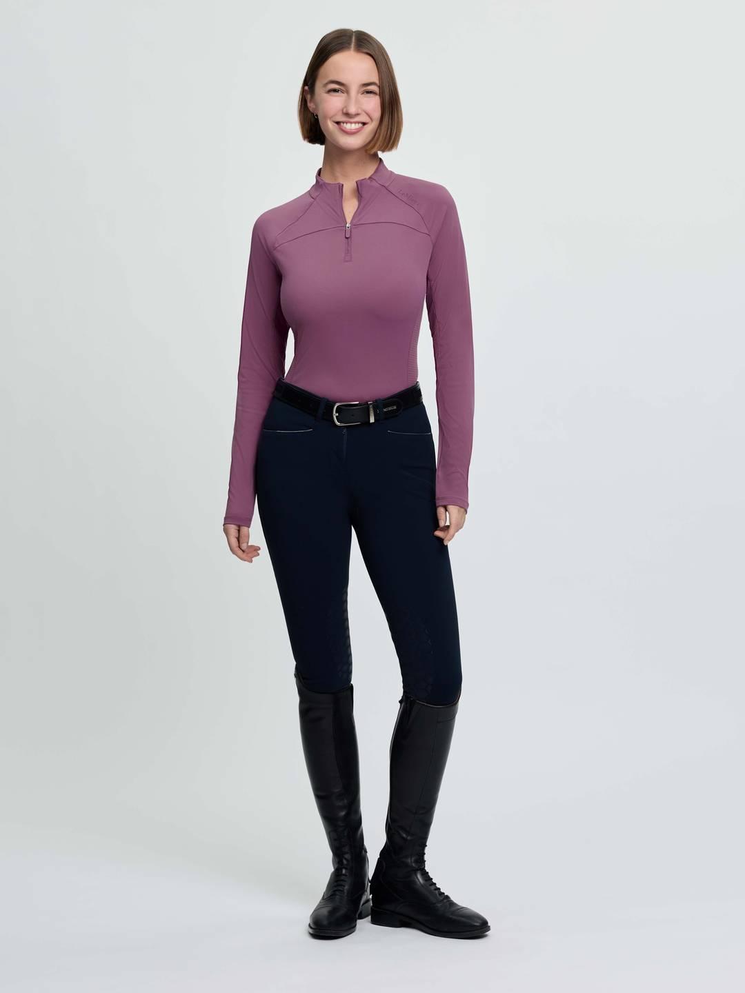 Full body front view of a female model wearing the purple LeMieux Airflow Long Sleeve Base Layer and dark colored breeches on a plain background