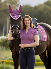 Front view of a female model wearing the purple LeMieux Lisa Mesh Short Sleeve Base Layer while standing in front of a dark colored horse