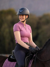 Angled front view of a female model wearing the purple LeMieux Lisa Mesh Short Sleeve Base Layer while riding a dark colored horse