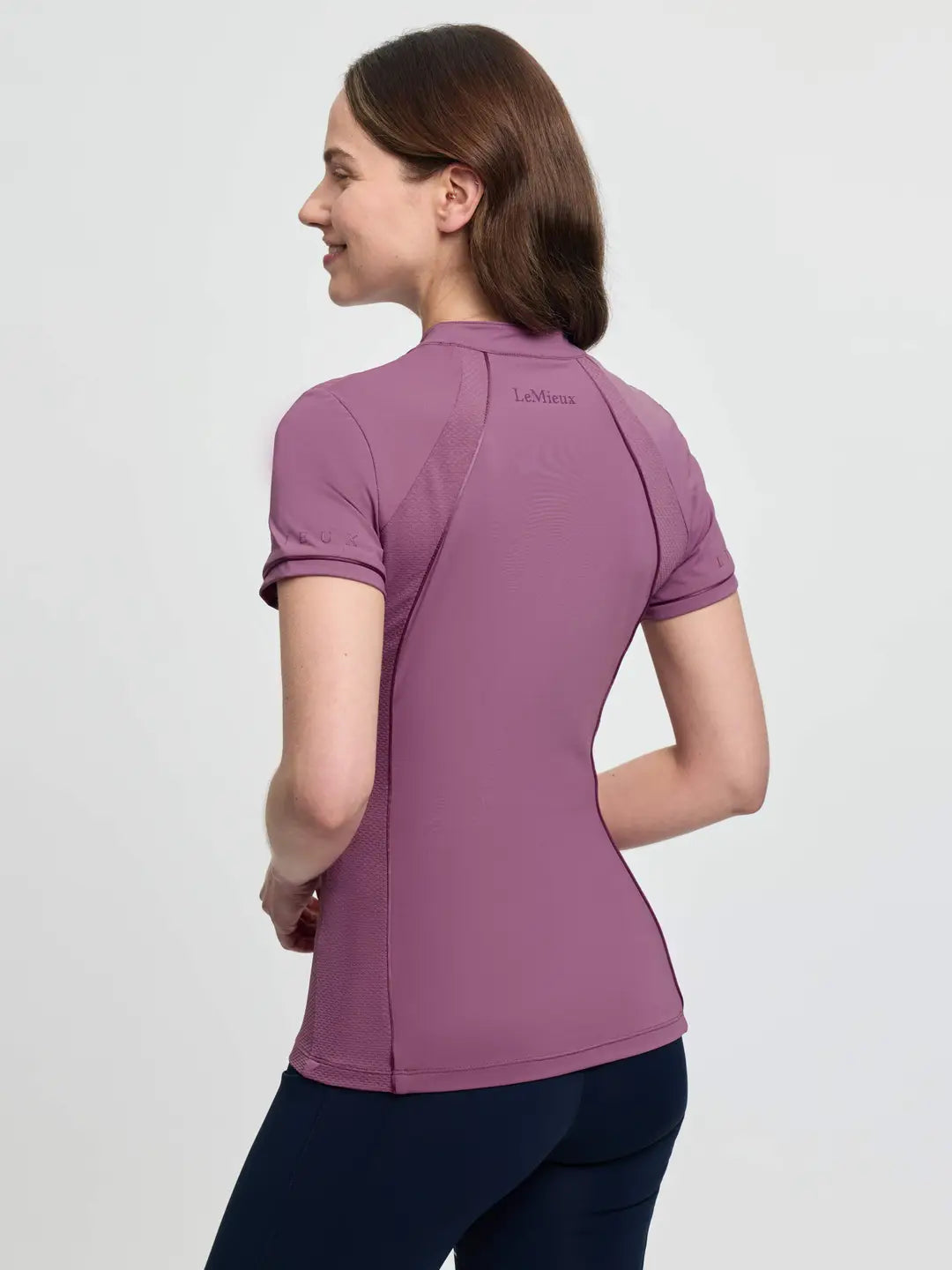 Angled back view of a female model wearing the purple LeMieux Lisa Mesh Short Sleeve Base Layer on a plain background