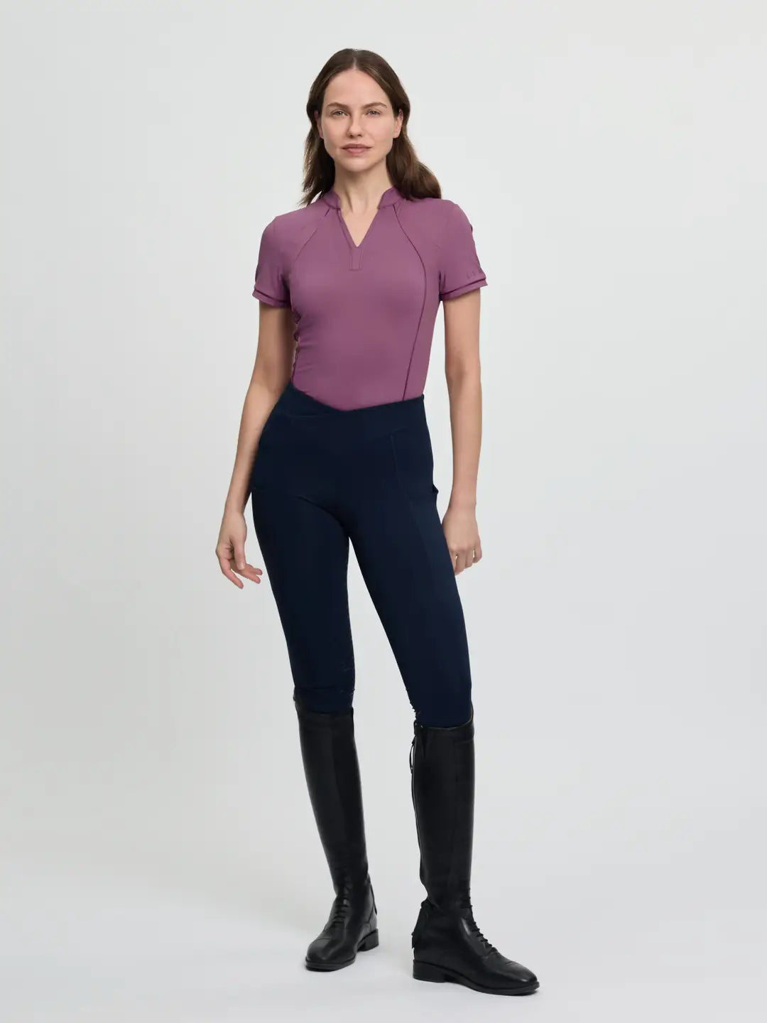 Full body front view of a female model wearing the purple LeMieux Lisa Mesh Short Sleeve Base Layer, navy tights, and black tall boots on a plain background