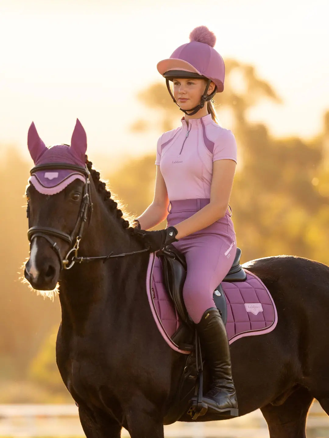 Front view of a female model wearing the dark pink LeMieux Classique Pom Hat Silk while riding a black horse