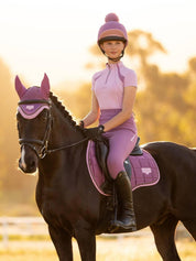 Angled front view of a young female model wearing the pink LeMieux Young Rider Mia Mesh Short Sleeve Base Layer while riding a dark colored horse outside