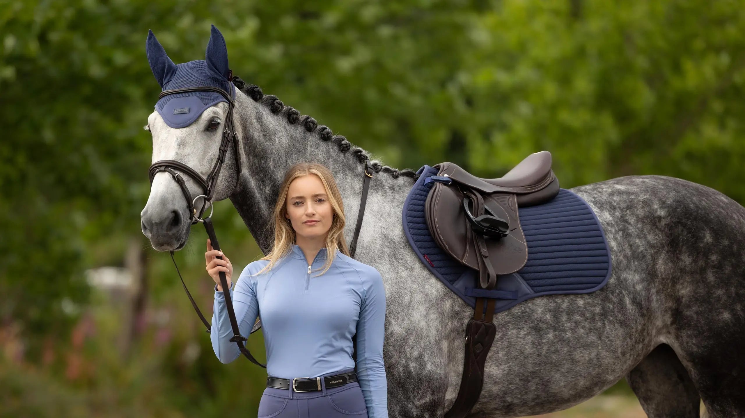 Woman standing next to a gray horse wearing the LeMieux Air-Tek Mesh Close Contact Square in deep blue under a brown saddle
