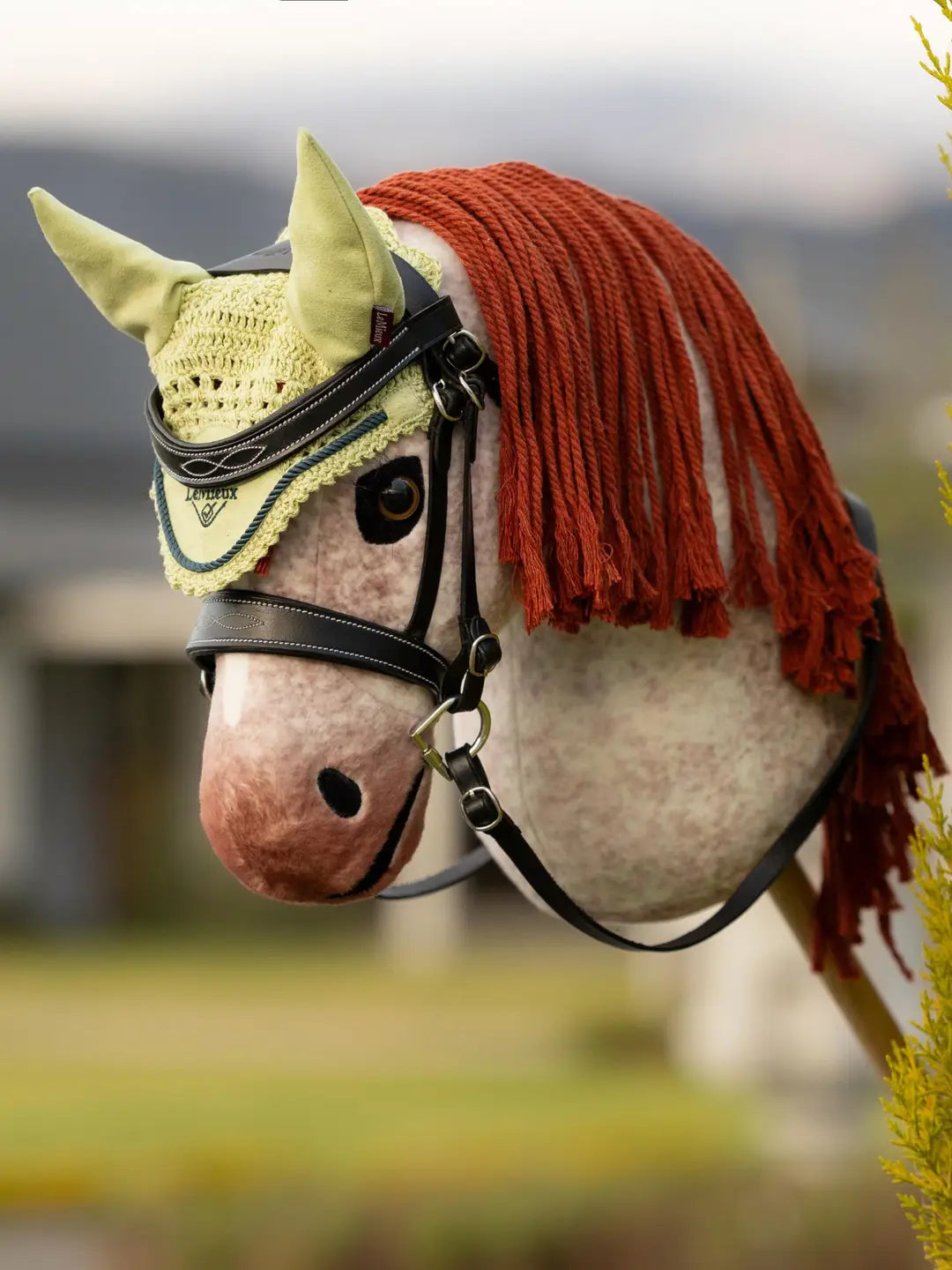 Angled front view of a red roan hobby horse wearing the green LeMieux Hobby Horse Ear Bonnet