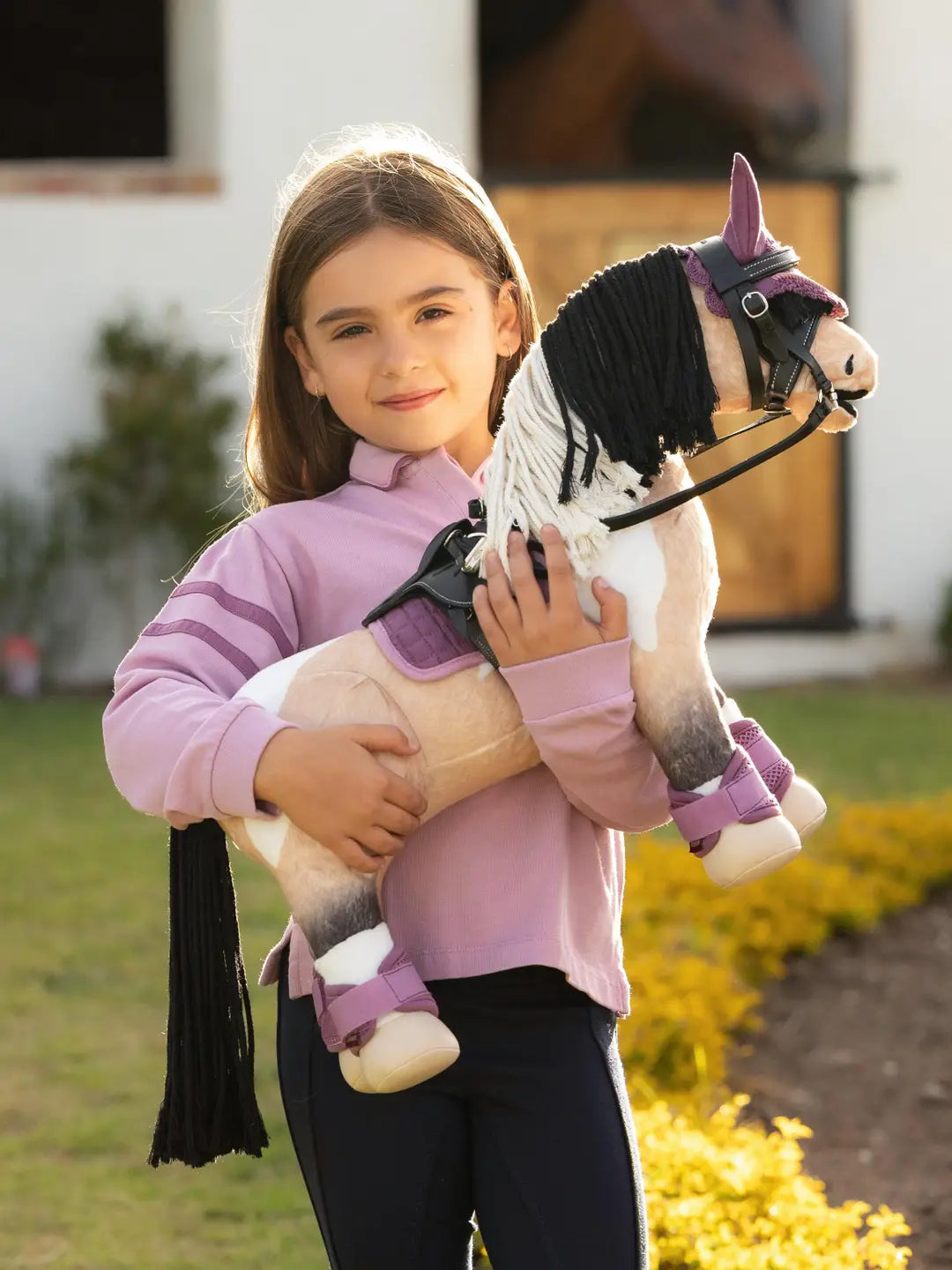 A young girl standing outside holding a buckskin pain stuffed animal horse - both dressed in purple