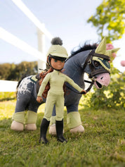 A doll dressed in green standing next to a gray appaloosa stuffed horse standing on grass