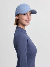 Side view of a female model wearing the powder blue LeMieux Lara Cap on a plain background