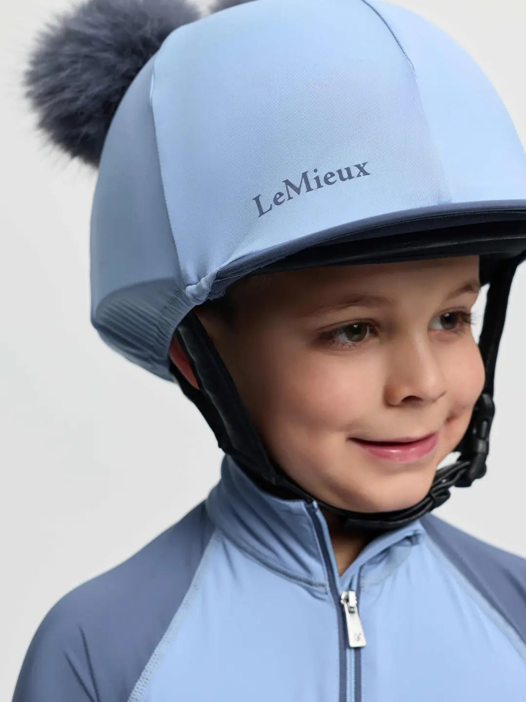 Close-up front view of a young male model wearing the Powder Blue LeMieux Mini Double Pom Hat Silk on a plain background