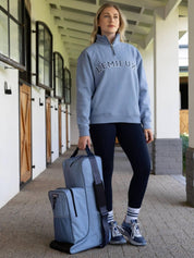 Front view of a female model holding the powder blue LeMieux Boot & Hat Bag while standing in a barn aisle