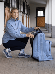 View of a female model putting tall boots in the powder blue LeMieux Boot & Hat Bag in a barn aisle