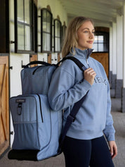 View of a female model wearing the powder blue LeMieux Boot & Hat Bag while standing in a barn aisle