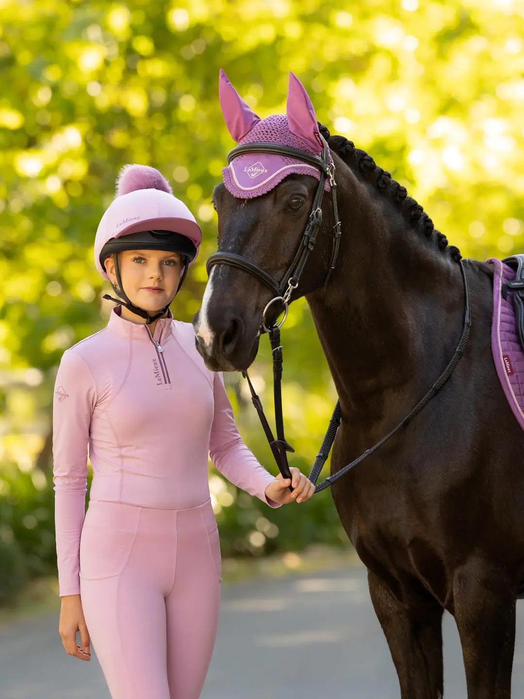 Front view of a female model wearing the pink LeMieux Classique Pom Hat Silk while standing outside next to a black horse