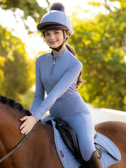 Front view of a young female model wearing the powder blue LeMieux Young Rider Classique Base Layer while sitting on a horse outside