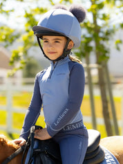 Side view of a young female model wearing the Powder Blue LeMieux Mini Double Pom Hat Silk while riding a pony outside