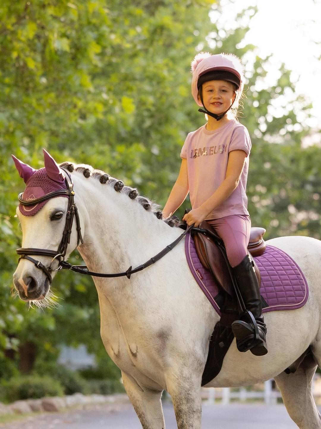 Angled front view of a female model wearing the fondant LeMieux Mini Alex T-Shirt while sitting on a gray pony outside