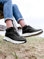 Side view of a model wearing a pair of the black LeMieux Kanta Waterproof Trainers at the beach