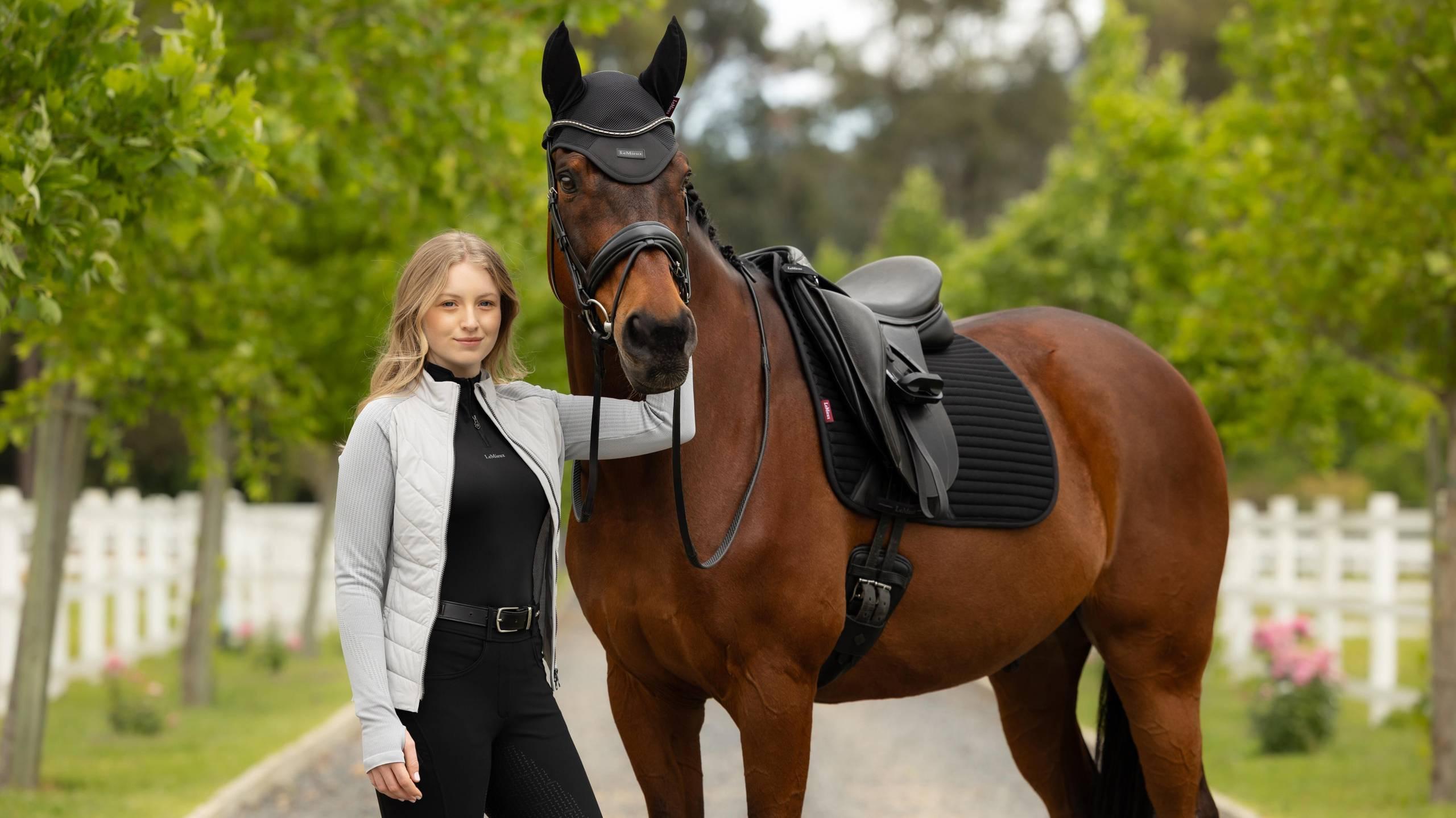Front view of a female model standing in front of a horse that is wearing the black LeMieux Air-Tek Mesh Dressage Square Pad while standing outside