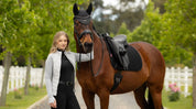 Front view of a female model standing in front of a horse that is wearing the black LeMieux Air-Tek Mesh Dressage Square Pad while standing outside