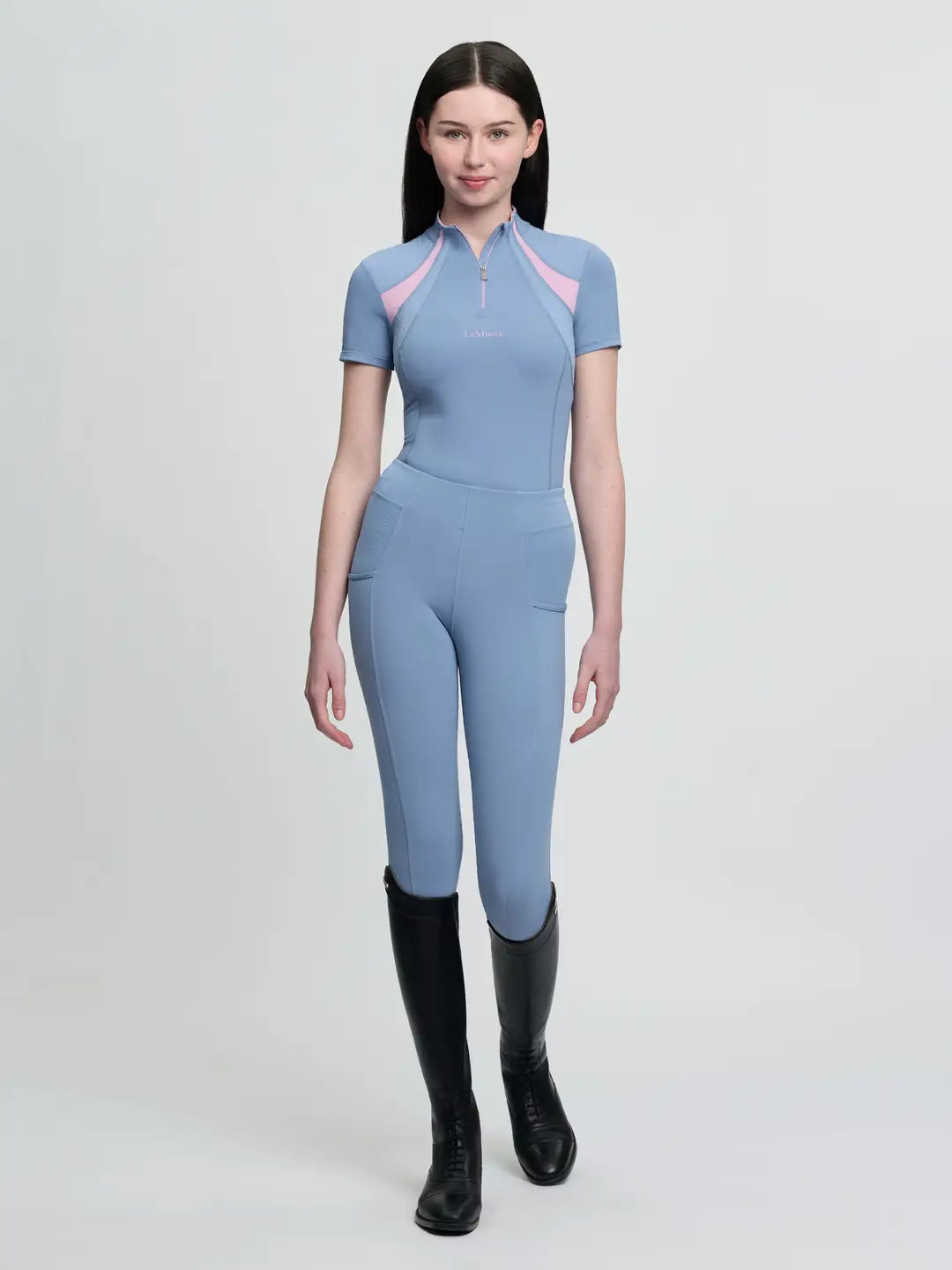 Full body front view of a young female model wearing the blue LeMieux Young Rider Mia Mesh Short Sleeve Base Layer with matching tights on a plain background
