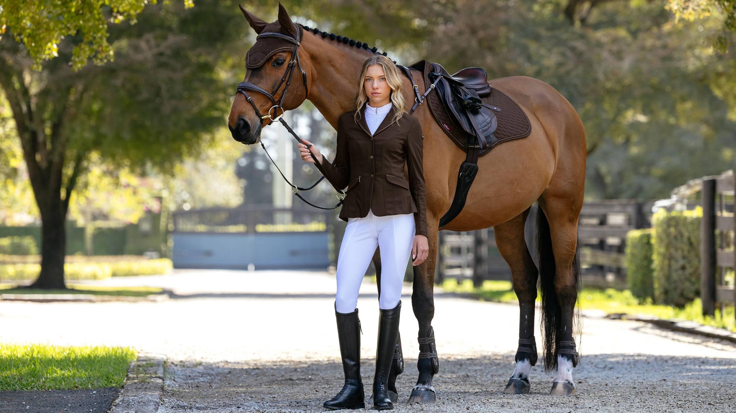 Angled front view of a female model standing in front of a bay horse wearing the espresso LeMieux Brilliance Close Contact Square Pad 