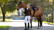 Angled front view of a female model standing in front of a bay horse wearing the espresso LeMieux Brilliance Close Contact Square Pad 
